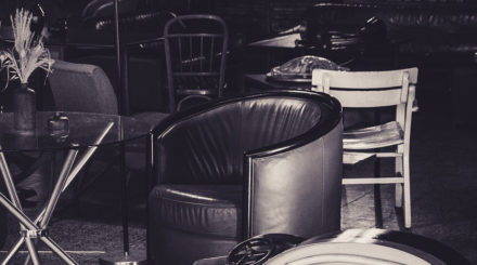 Et svart-hvitt bilde av en eldre stol og andre brukte møbler i bakgrunnen.
